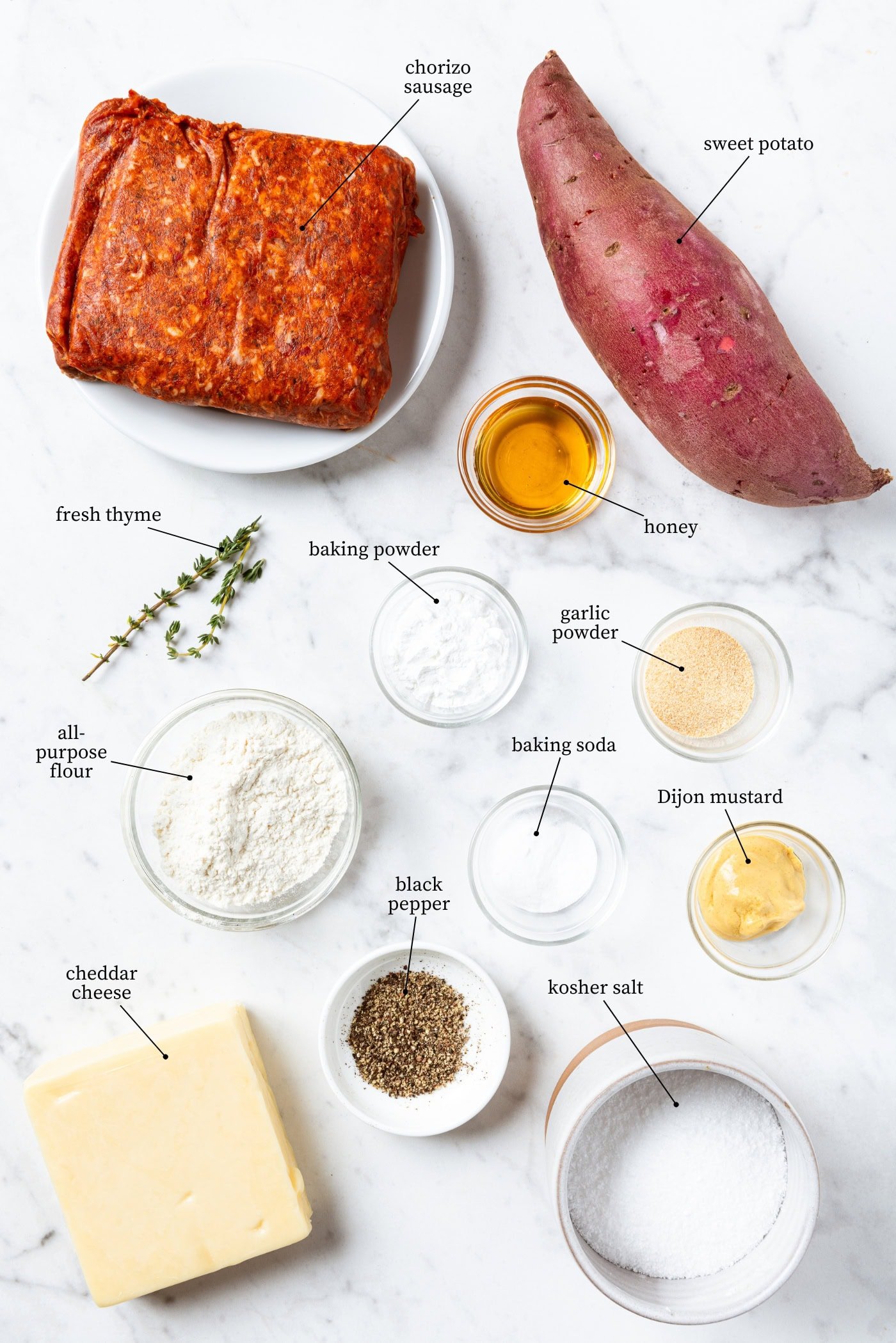 White marble surface filled with ingredients needed to make sweet potato puffs including sausage, sweet potato, flour, spices, cheese, and more.