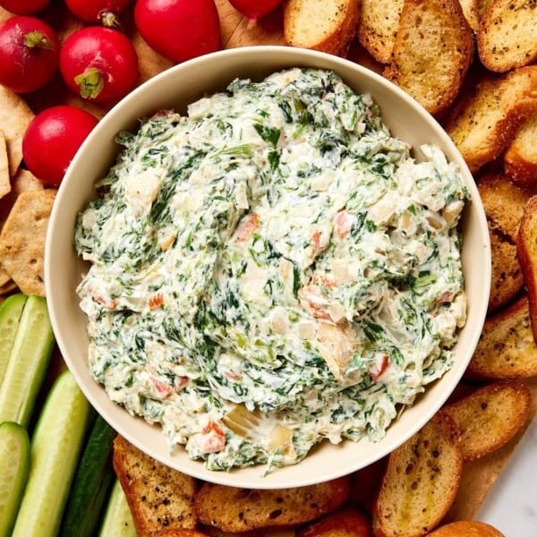 White bowl with spinach artichoke dip all on wooden cutting board with fresh vegetables all around.