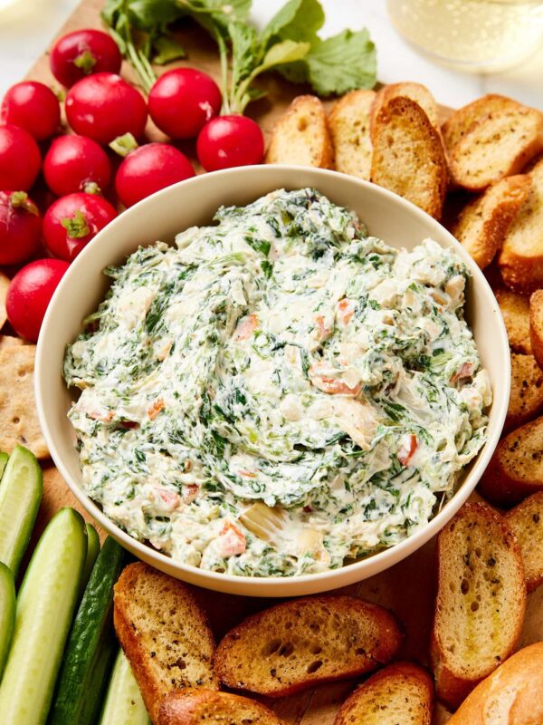 White bowl filled with creamy spinach artichoke dip with crostini and vegetables all around on a wooden cutting board.