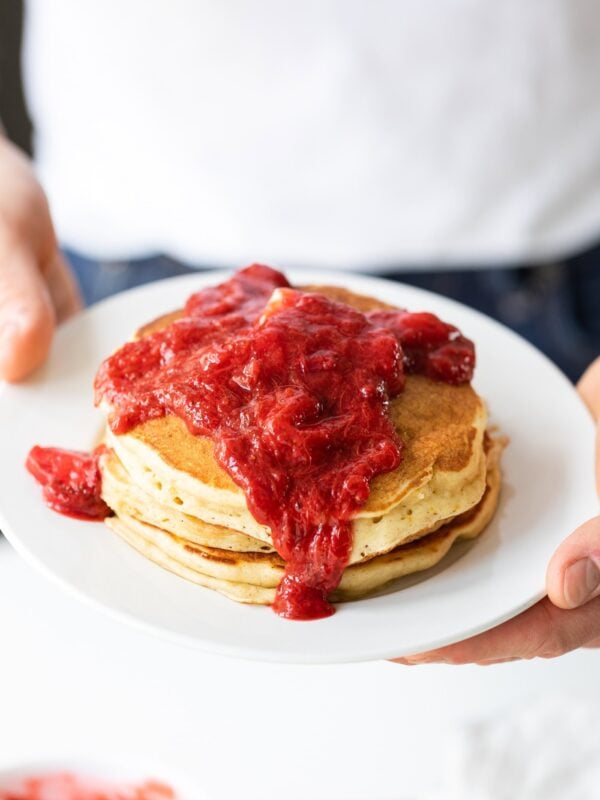 White plate with red-colored strawberry rhubarb sauce spilling over golden pancakes.