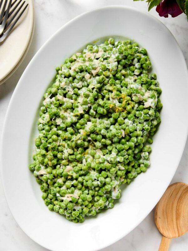 White marble surface with white platter filled with brightly colored creamed peas with spoon to the side.