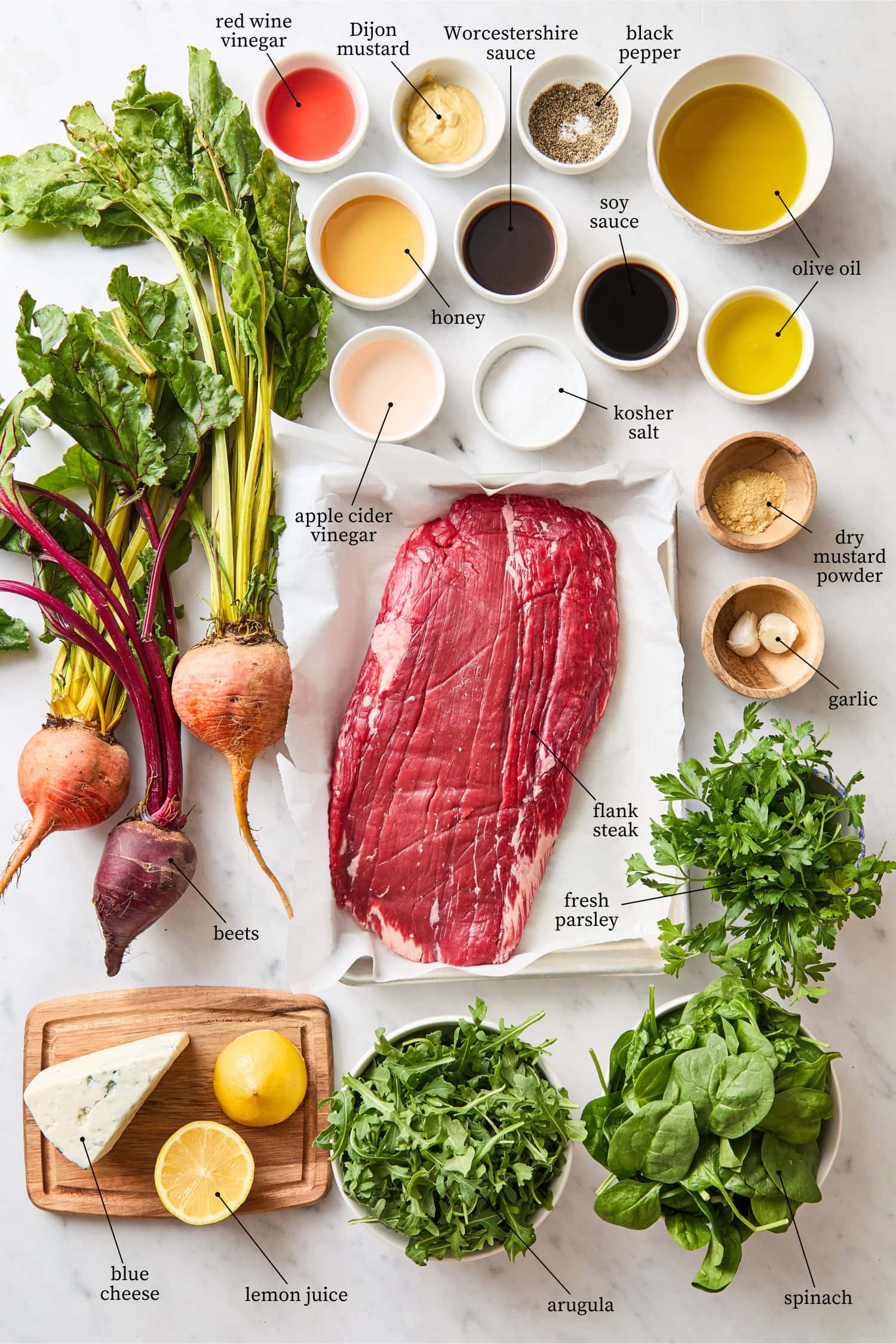 White marble surface with ingredients needed for steak and beet salad including flank steak, beets, herbs, arugula, and more.