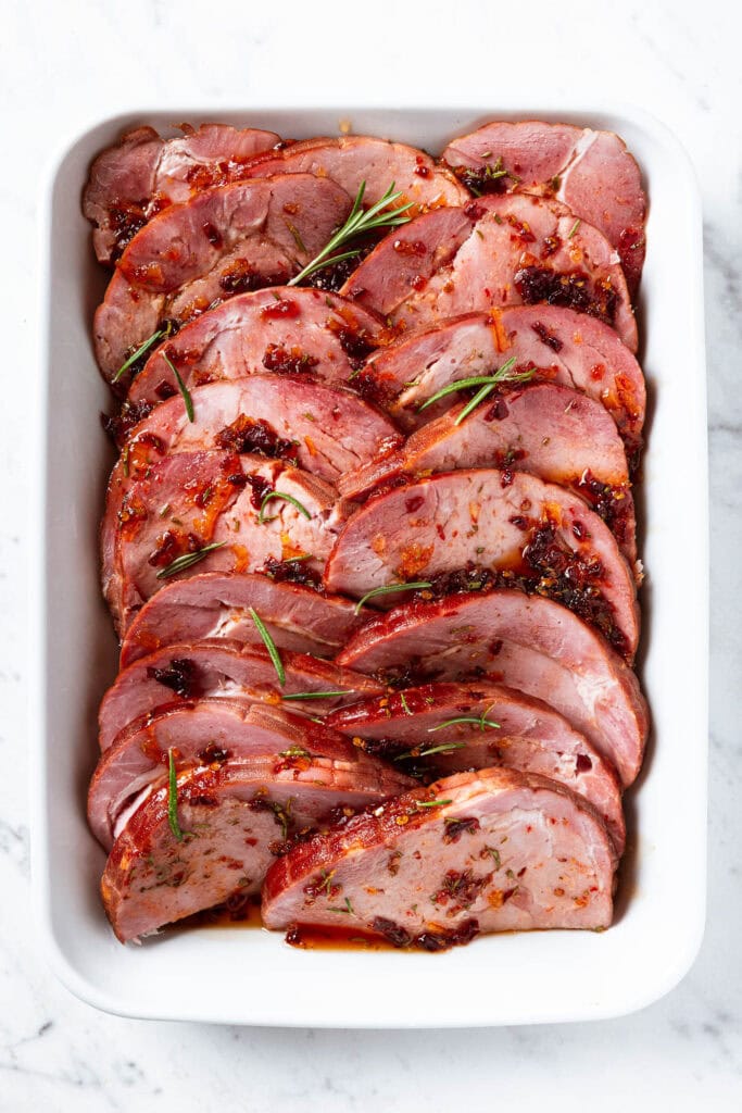 Sliced ham covered with orange chipotle glaze and rosemary in a baking dish.