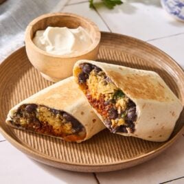 Brown plate with breakfast burrito sliced in half with bowl of sour cream in background.