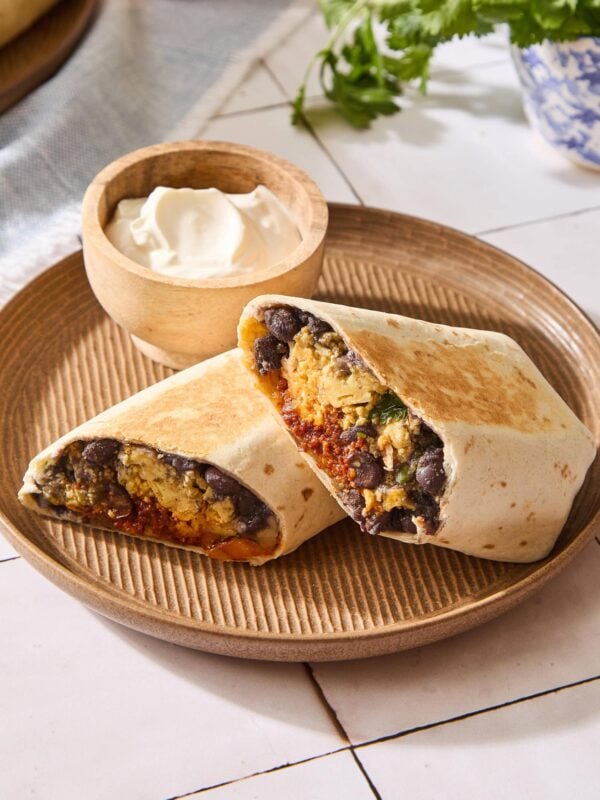 Brown plate with breakfast burrito sliced in half with bowl of sour cream in background.