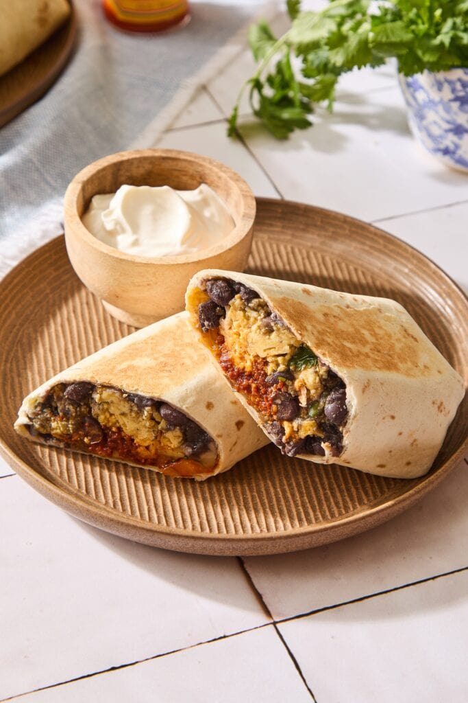 Brown plate with breakfast burrito sliced in half with bowl of sour cream in background.