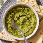 White bowl filled with swirled cilantro pesto with pita bread all around.