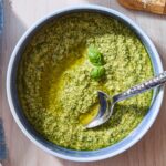 Blue bowl filled with green colored basil pesto with spoon resting in bowl.