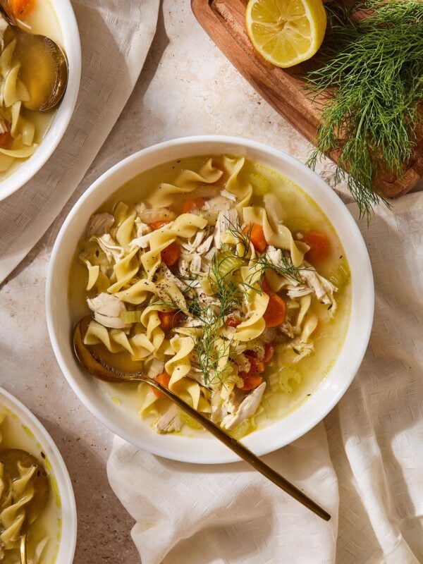 Bowl filled with homemade chicken noodle soup with spoon resting in bowl with carrots and fresh herbs on top.