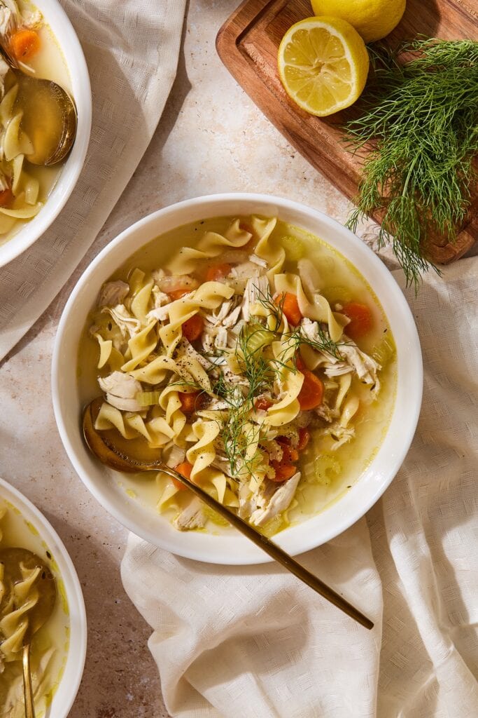 Bowl filled with homemade chicken noodle soup with spoon resting in bowl with carrots and fresh herbs on top.