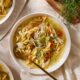 Bowl filled with homemade chicken noodle soup with spoon resting in bowl with carrots and fresh herbs on top.