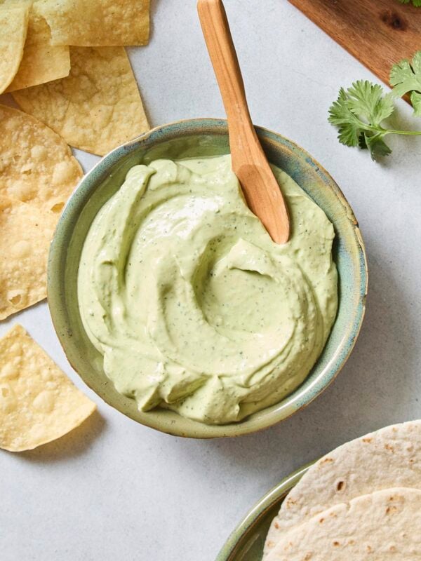 Green bowl with smooth avocado crema sauce with wooden spoon and tortilla chips.