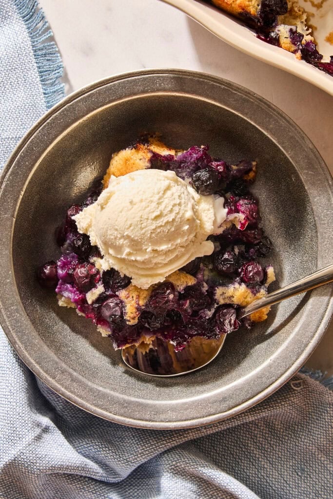 Metal bowl with serving of blueberry cobbler with scoop of ice cream on top.