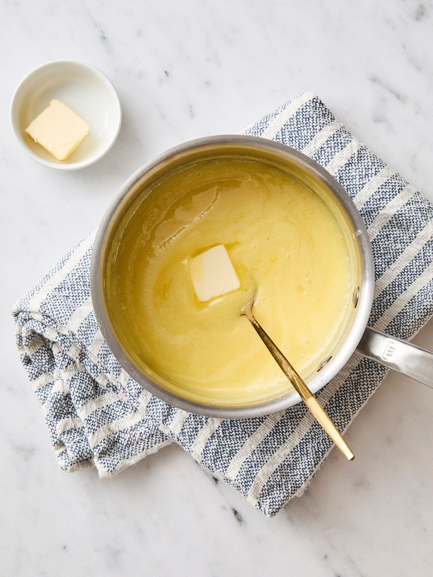 White marble with saucepan filled with lemon curd with butter being stirred in.