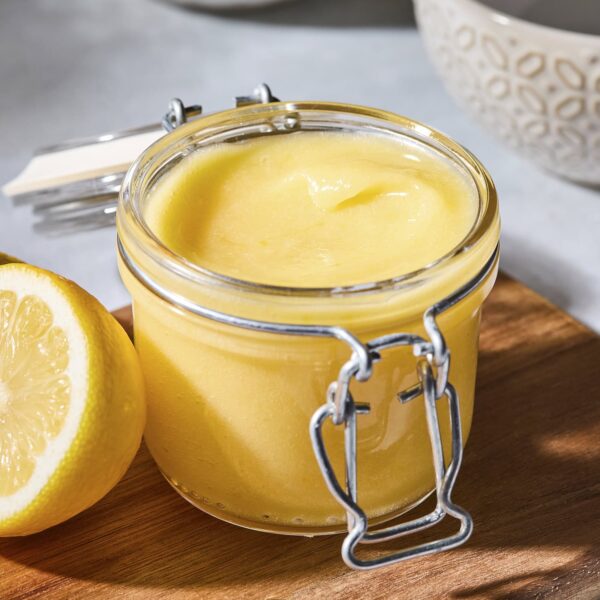 Glass jar filled with lemon curd sitting on a cutting board with fresh lemons.