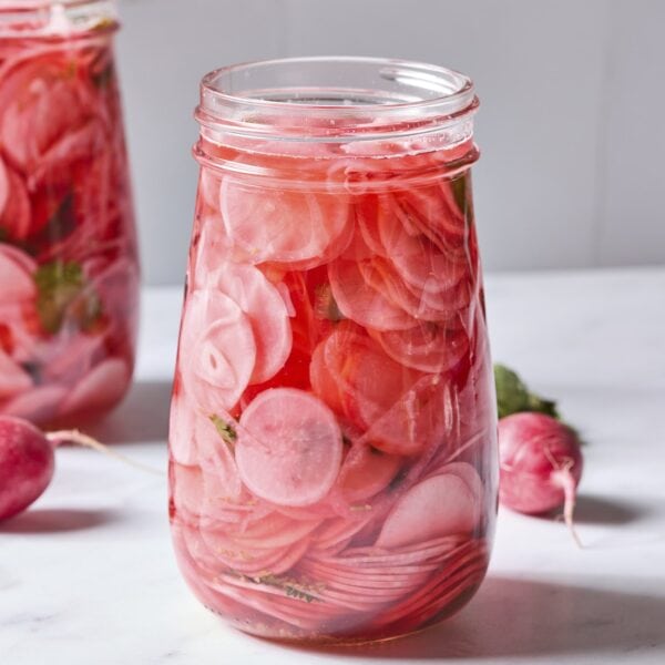 Glass jar with pink liquid and quick pickled radishes with more radishes and jar to the side.