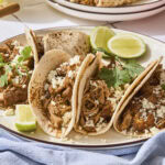 Four chicken tacos with cheese and cilantro on top sitting on a plate.