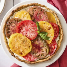 Tile surface with baked tomato tart with slices of tomato shingled on top garnished with fresh basil.