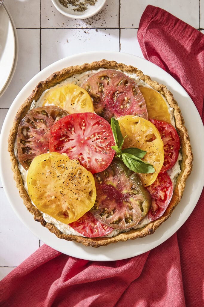 Tile surface with baked tomato tart with slices of tomato shingled on top garnished with fresh basil.