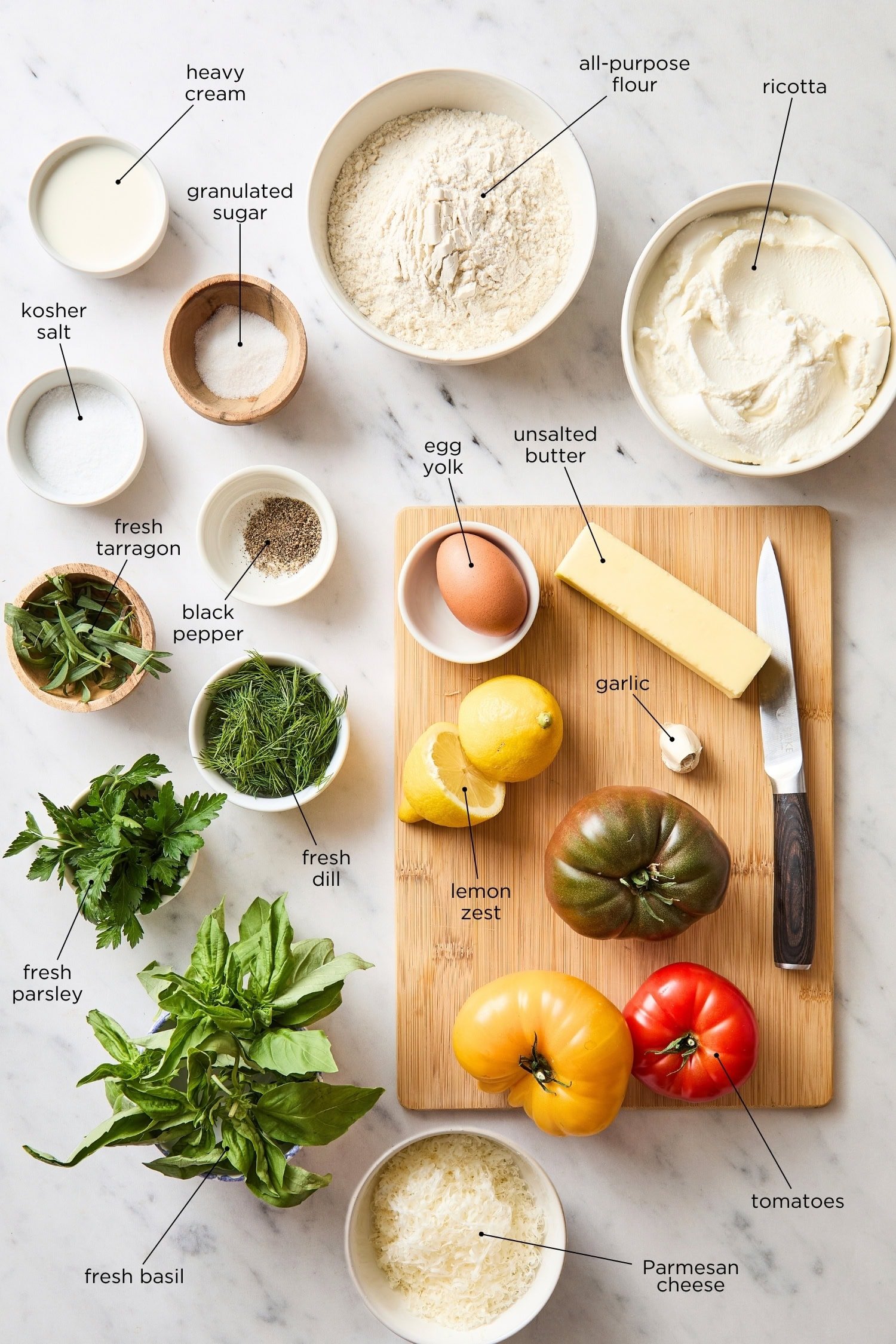 Marble surface with ingredients needed for tomato tart including ricotta, tomatoes, fresh herbs, and more.