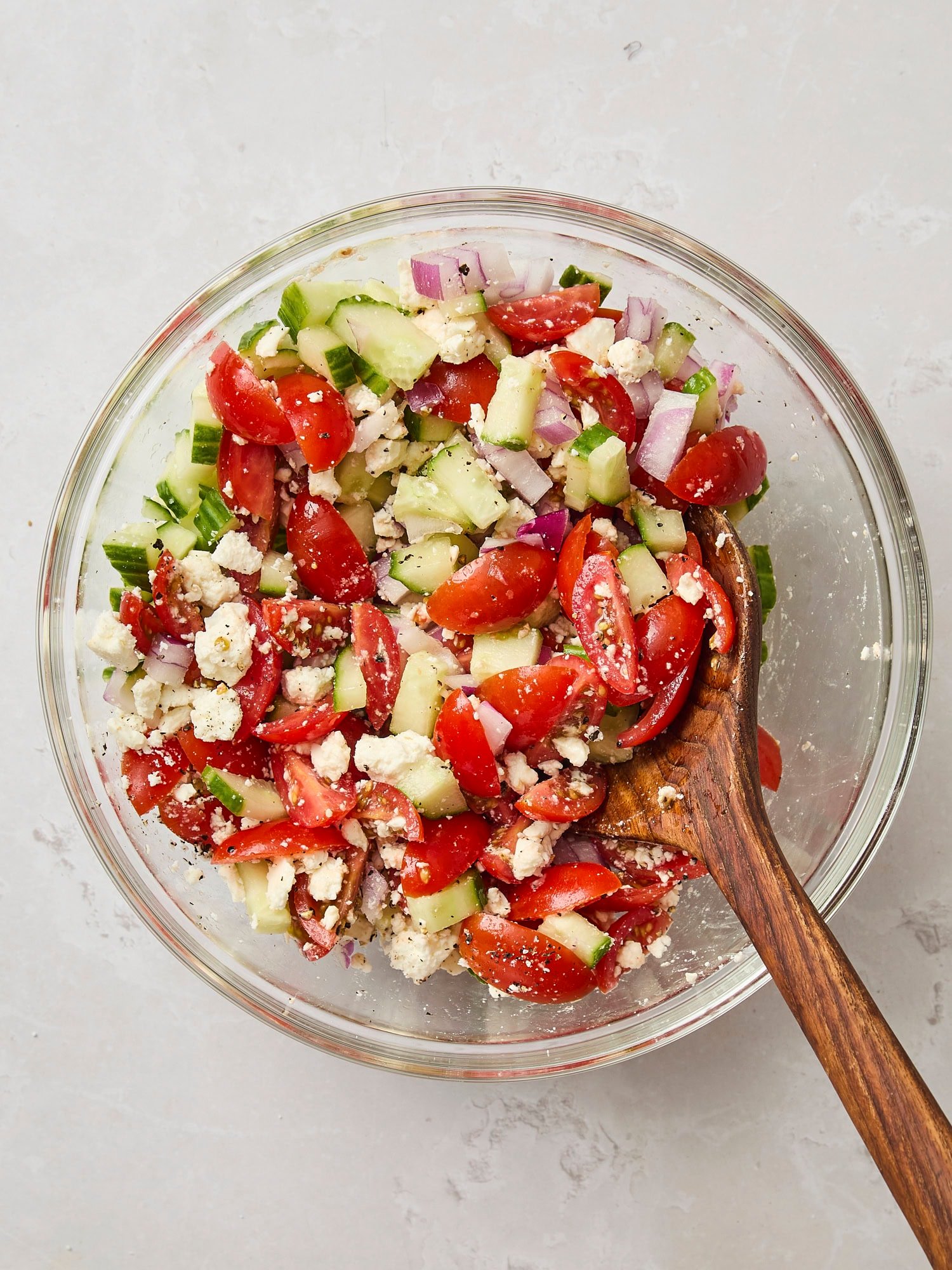 Glass bowl with Greek salad being stirred with wooden spoon.