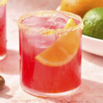 Glass filled with pink colored margarita with orange slice.