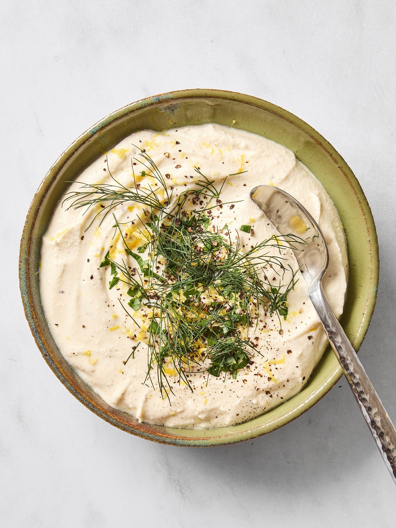 Bowl with feta dip topped with lemon zest, herbs, and olive oil with spoon.