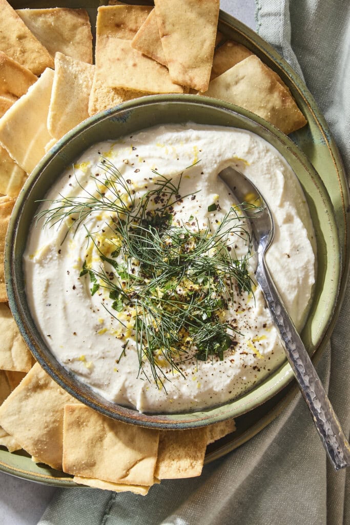 Whipped feta dip spread into a bowl topped with herbs and lemon zest with pita chips around.