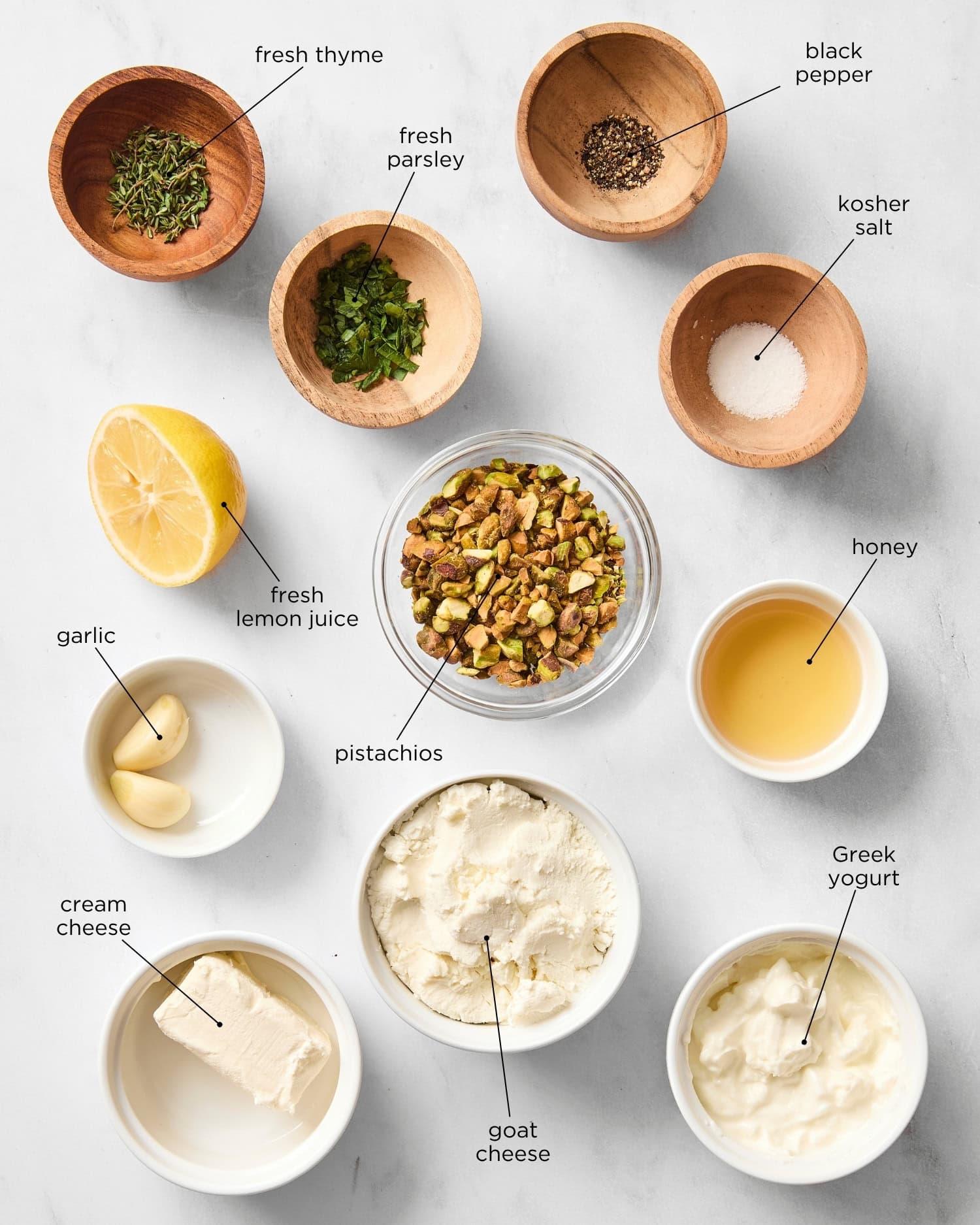 Marble surface with ingredients to make goat cheese dip including pistachios, herbs, honey, cream cheese, goat cheese, and more.
