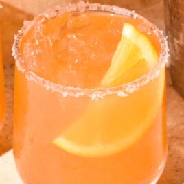 Coral-colored margarita sitting on a stone surface with an orange wedge in the drink.