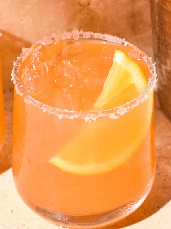 Coral-colored margarita sitting on a stone surface with an orange wedge in the drink.
