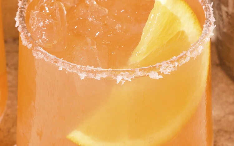 Coral-colored margarita sitting on a stone surface with an orange wedge in the drink.