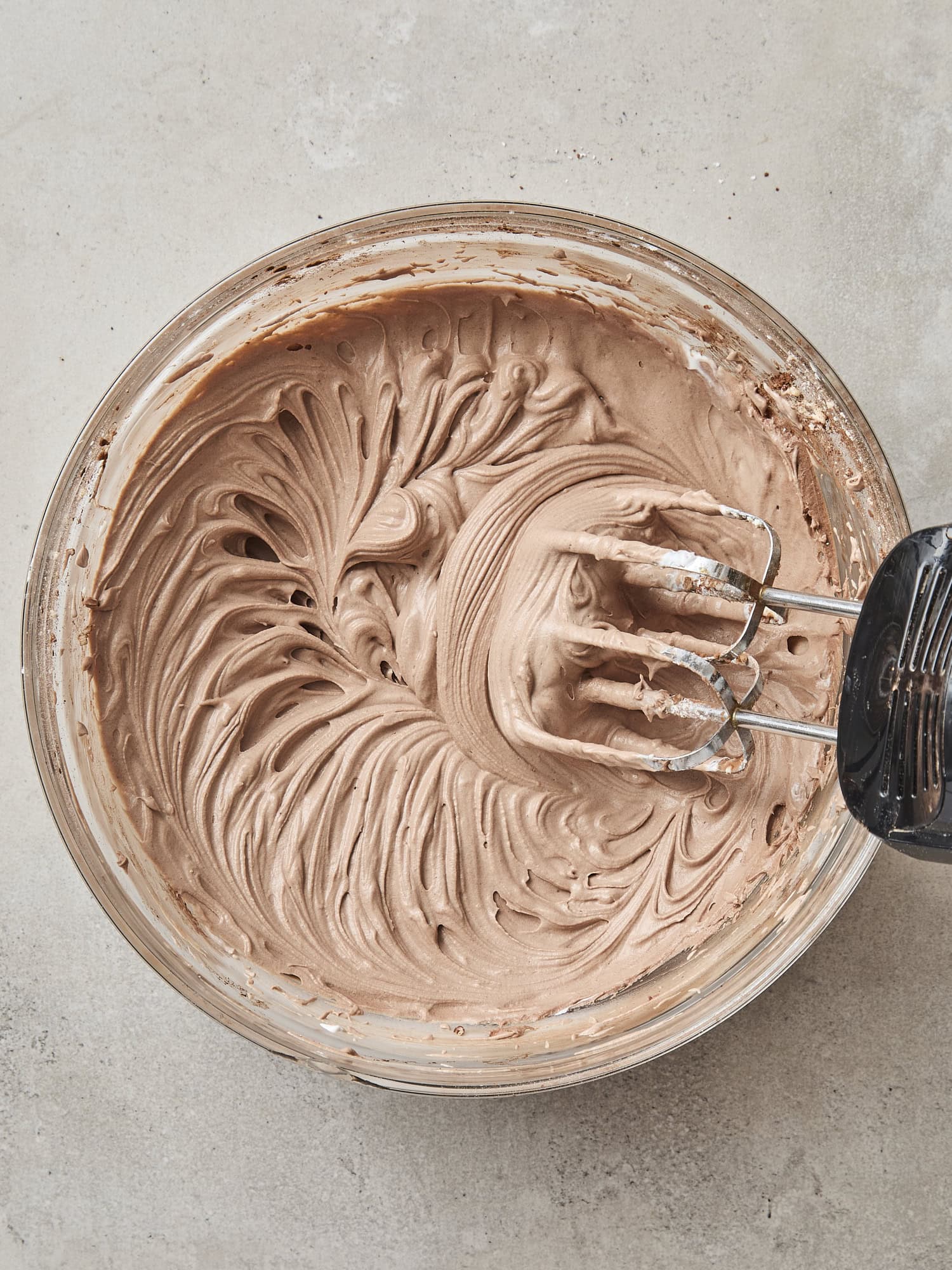 Glass bowl filled with swirled chocolate frosting with mixer mixing everything together.