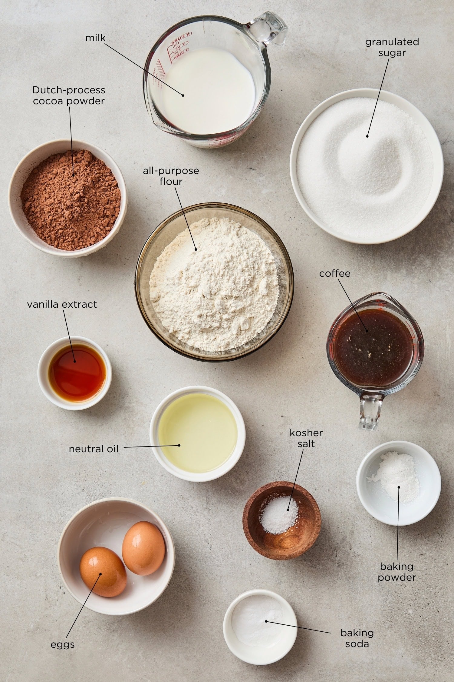 Concrete surface with ingredients for chocolate cake including sugar, cocoa, oil, eggs, and more.