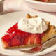 Piece of strawberry pie on a white plate with dollop of whipped cream on top.
