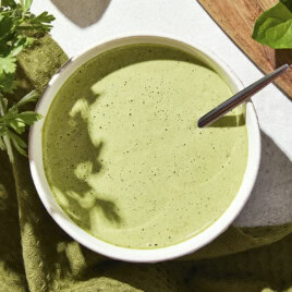 Small bowl filled with green colored dressing with a cutting board and more herbs to the side.