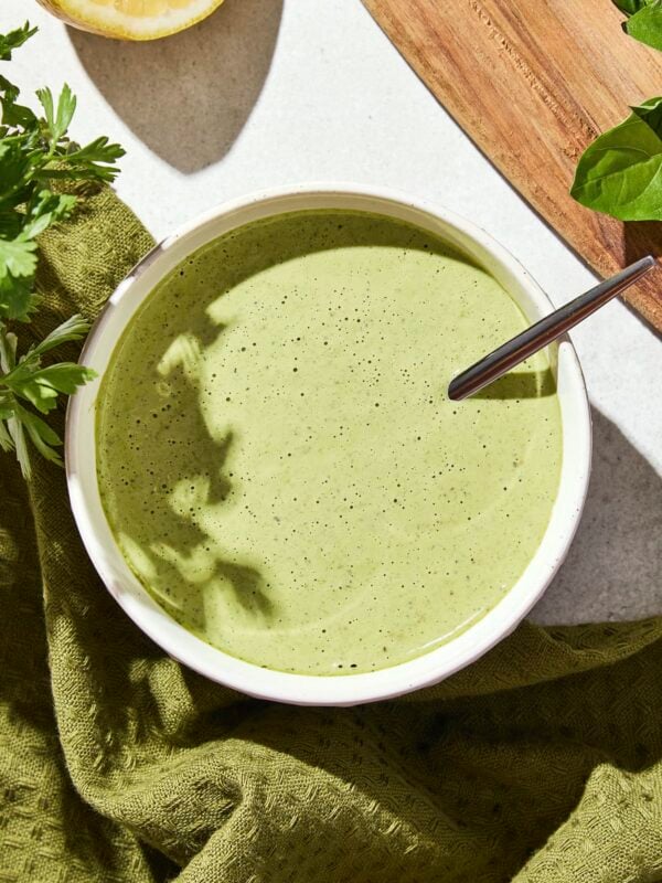 Small bowl filled with green colored dressing with a cutting board and more herbs to the side.