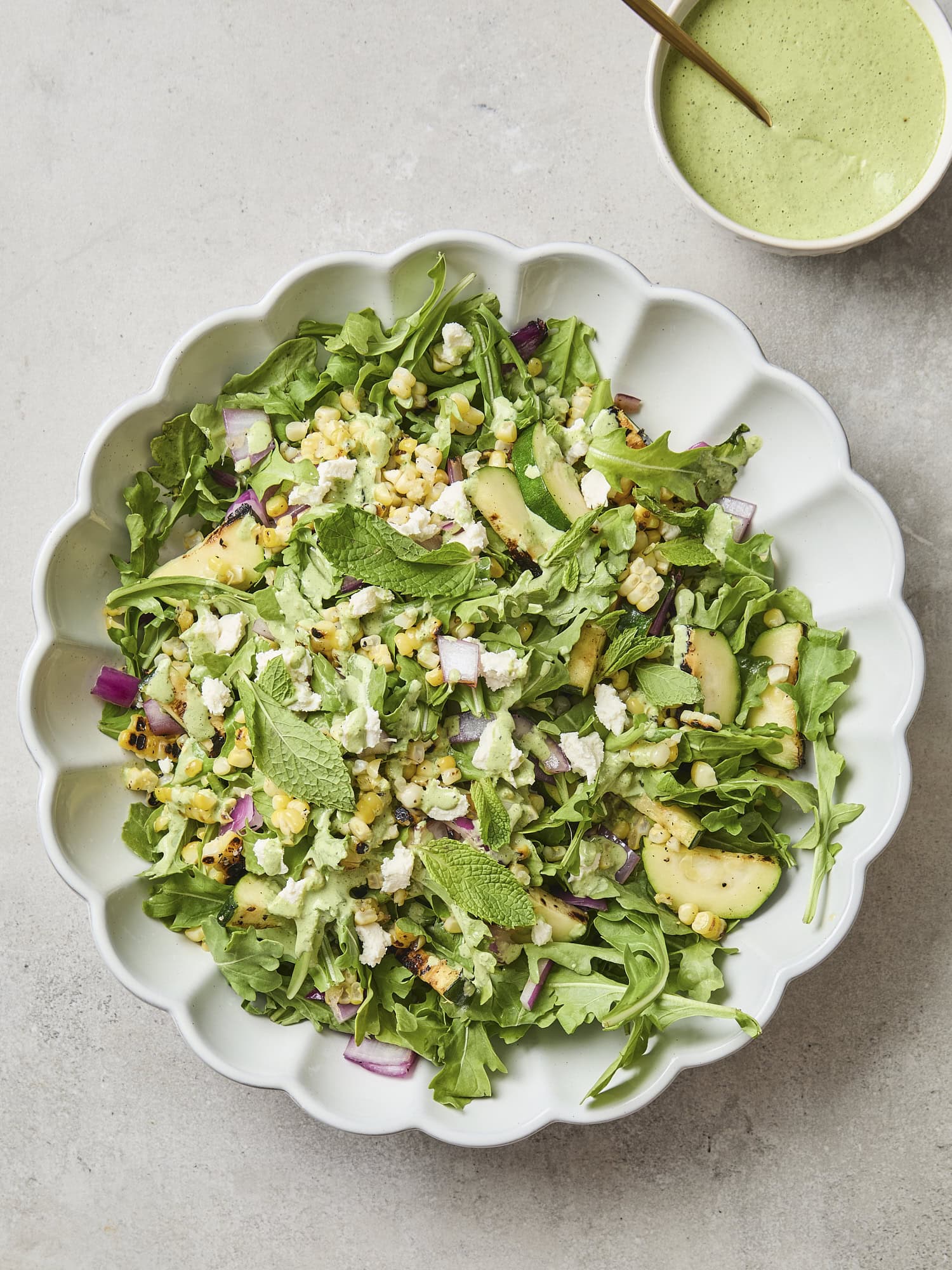 White bowl filled with salad with sweet corn, red onion, zucchini, and dressing with more dressing to the side.