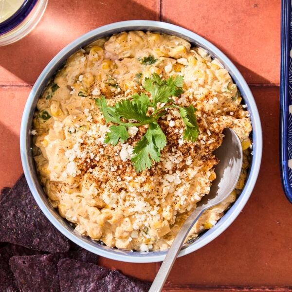 Bowl with spoon scooping out creamy street corn dip on red tile surface.