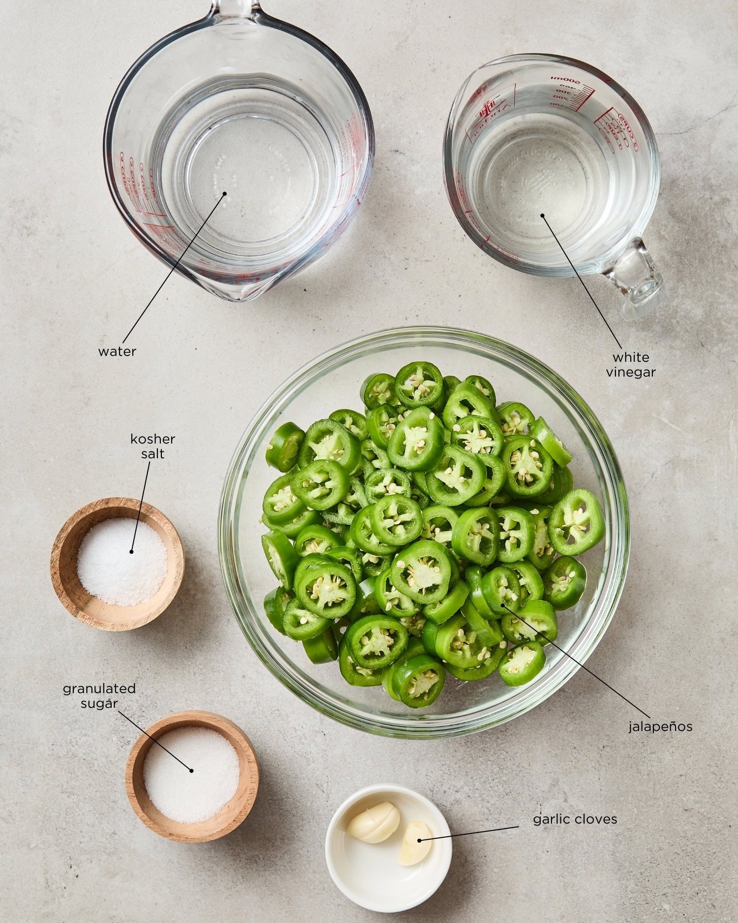 Ingredients needed to make quick-pickled jalapeños sit on a counter, including vinegar, water, sugar, kosher salt, jalapeños, and garlic.