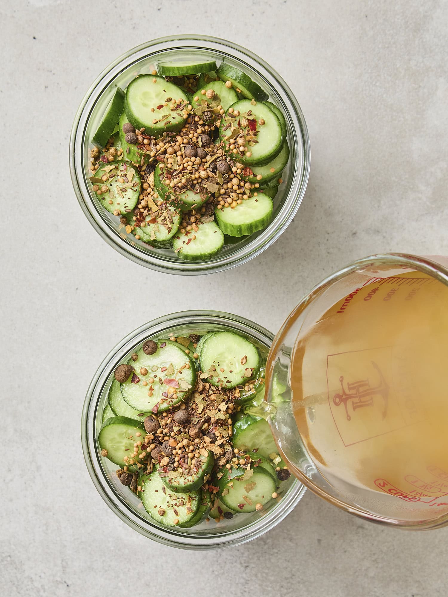 Measuring cup pouring apple cider vinegar brine on top of sliced cucumbers.