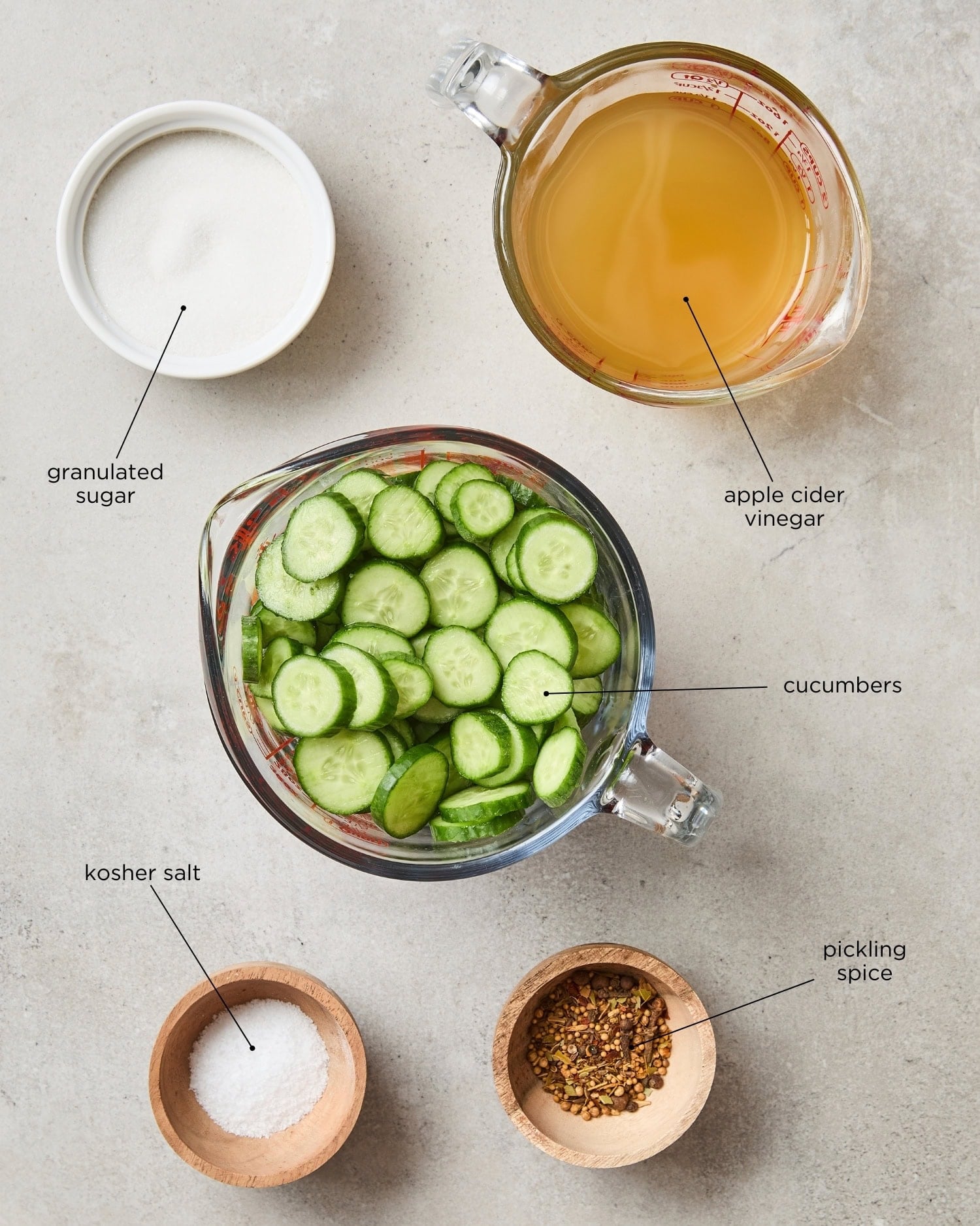 Concrete surface with ingredients for sweet pickles including cucumbers, apple cider vinegar, spices, and more.