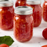 A jar of preserved diced tomatoes rests on a marble counter next to tomatoes.