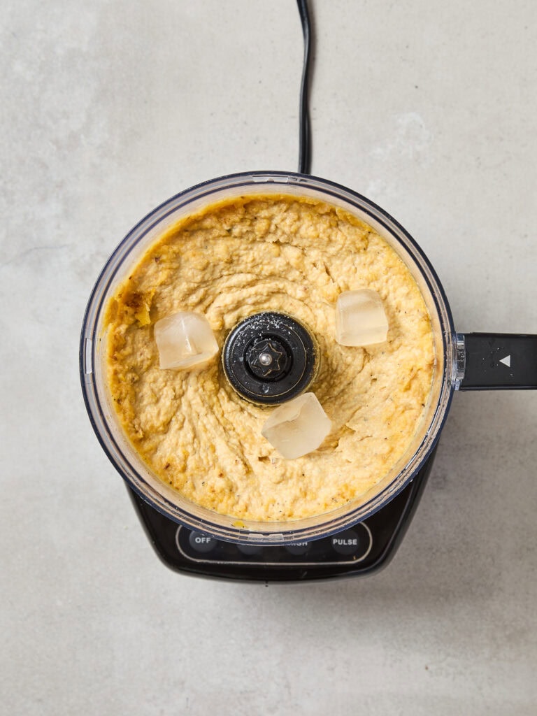 Food processor with squash hummus with three ice cubes sitting on surface.