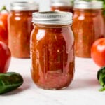 Jars of canned homemade salsa sitting on marble surface with tomatoes and peppers around.