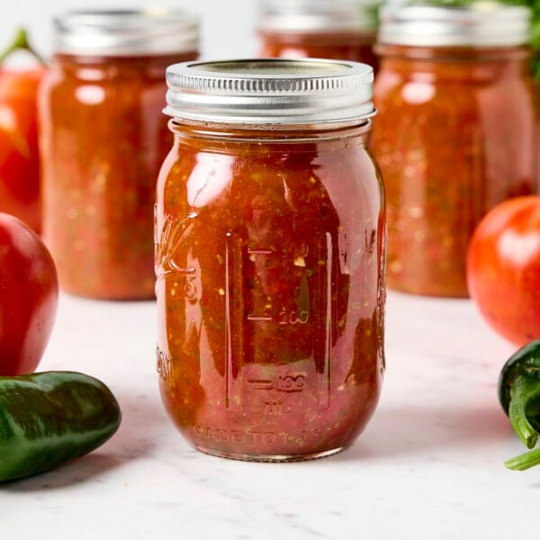 Jars of canned homemade salsa sitting on marble surface with tomatoes and peppers around.