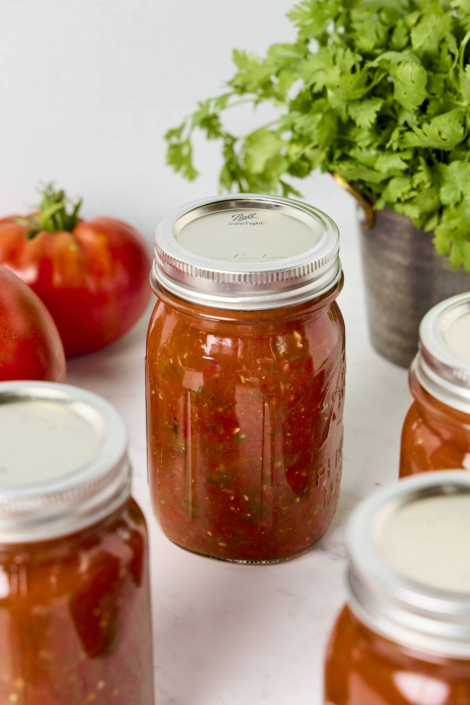 Jar of canned salsa with more jars around with cilantro and tomatoes in the background.