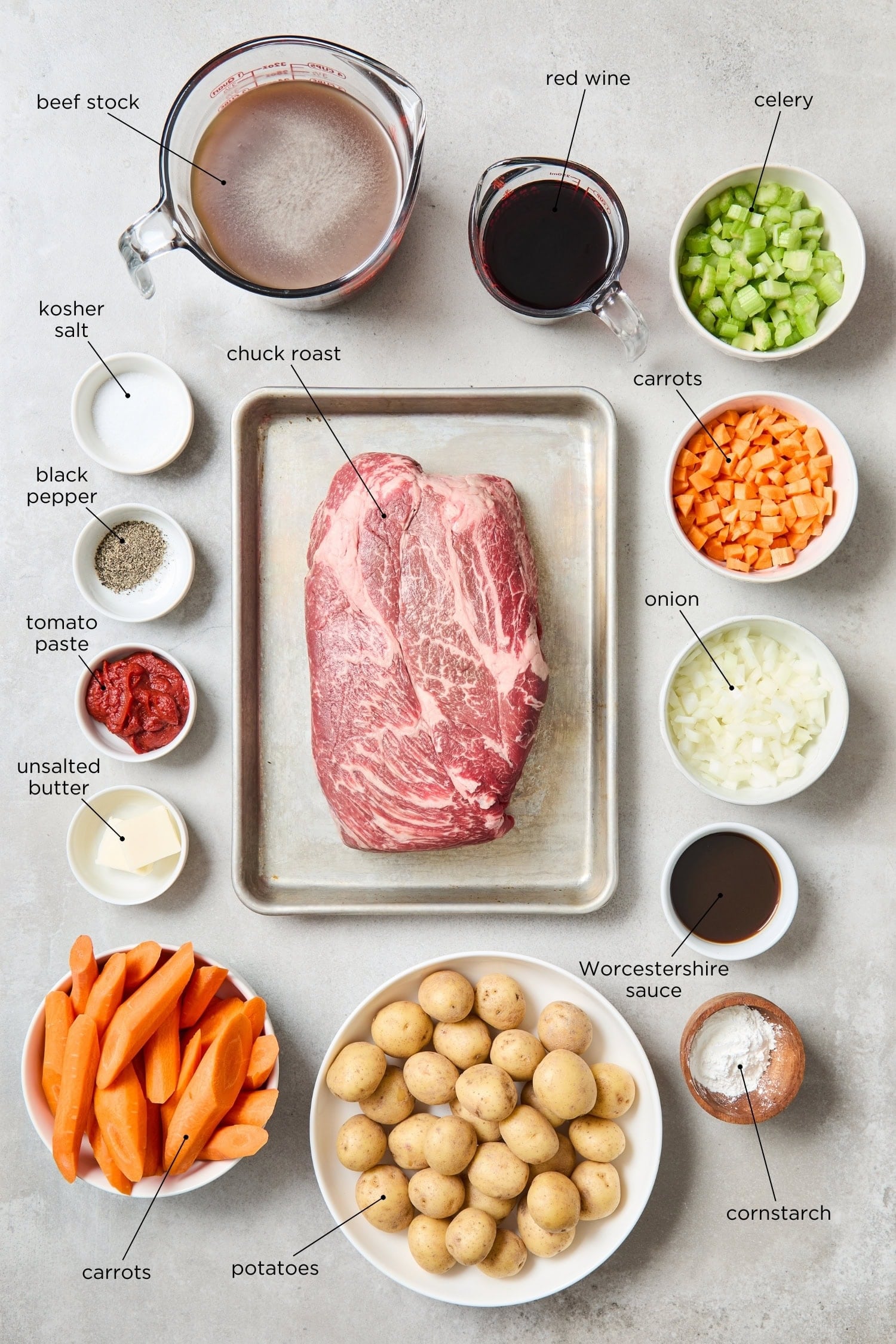 Concrete surface with ingredients needed for classic pot roast recipe including beef, carrots, potatoes, celery, stock, and more.