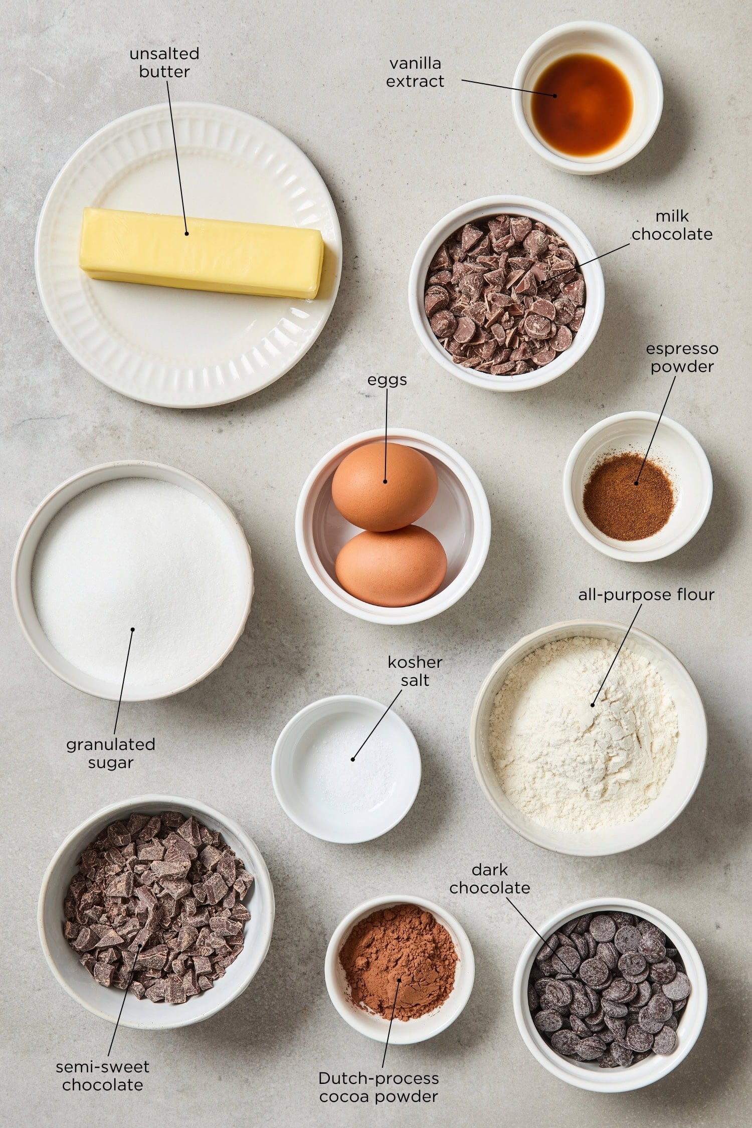 Concrete surface with ingredients needed for brownies including three types of chocolate, flour, eggs, and more.