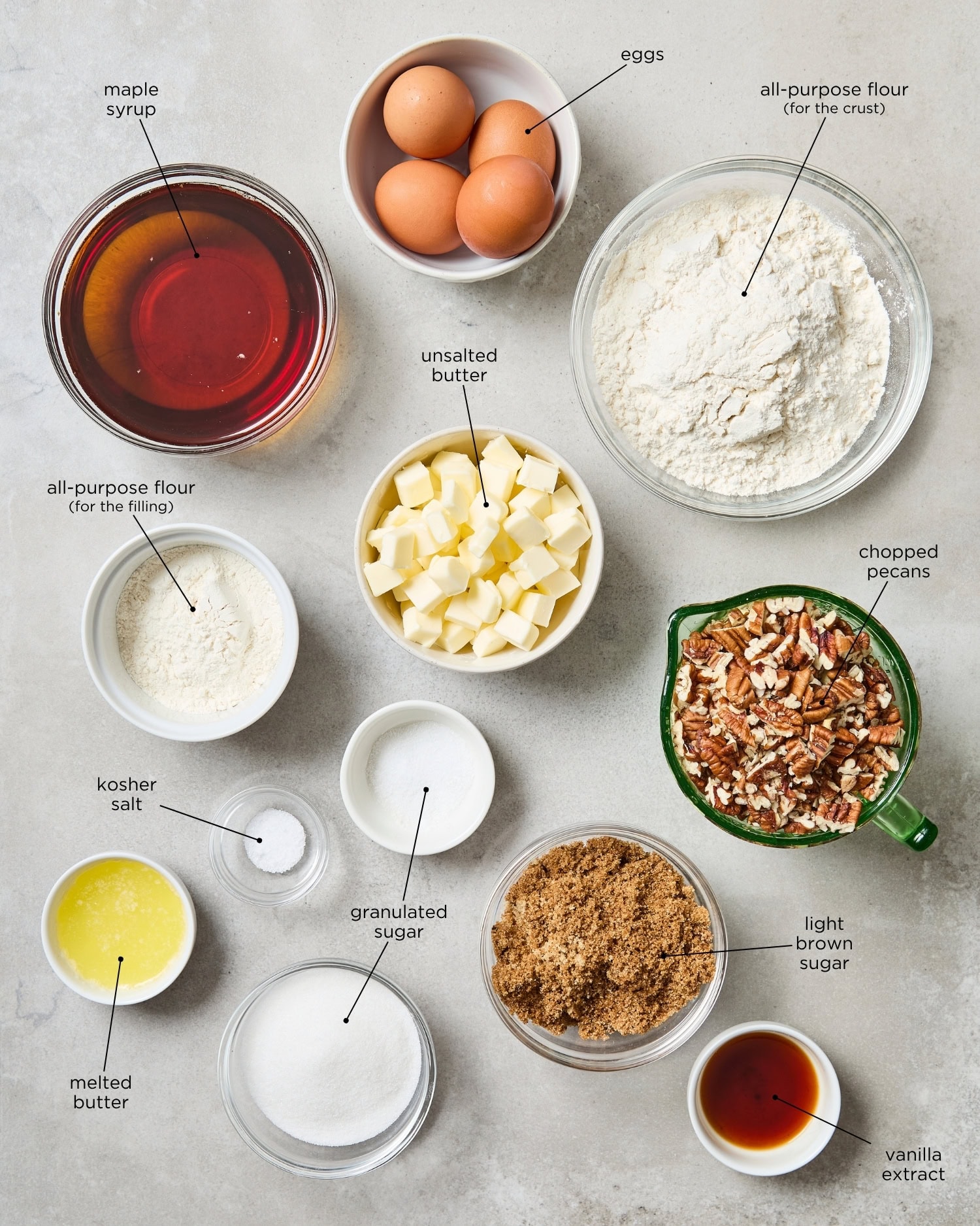 Concrete surface with ingredients for pecan pie bars including maple syrup, chopped pecans, flour, sugar, and more.